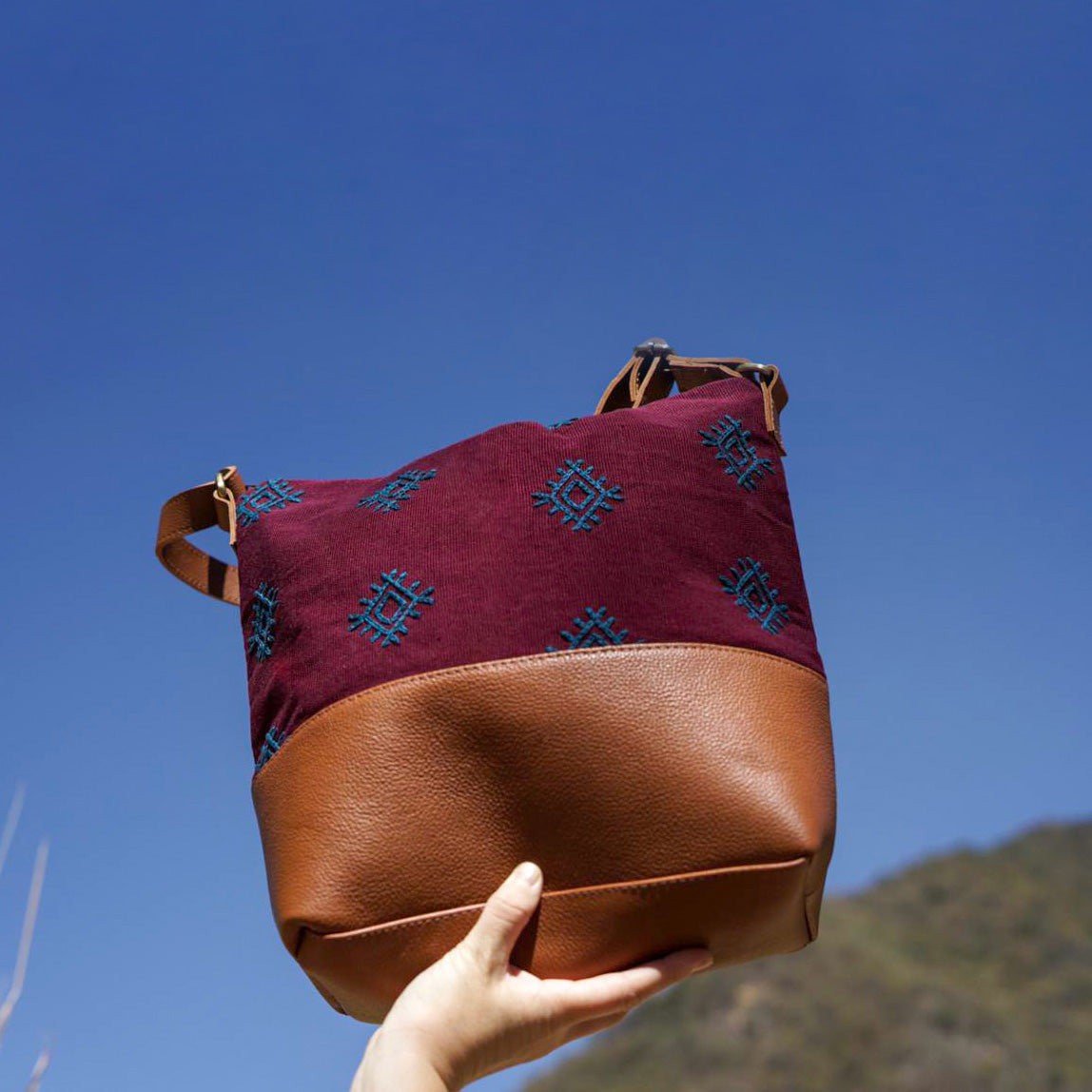 A model holds the hand woven artisan Lidia Hobo Shoulder Bag in Mountain Dusk. The photo is taken outside near a grassy hill.