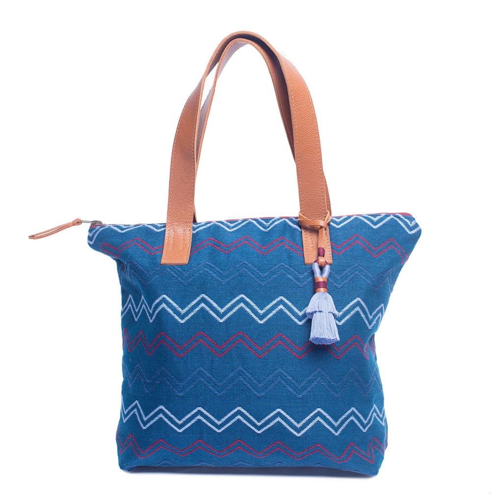 Front of the Angela Handwoven Tote in Lake Ripple pattern. It has horizontal zigzag stripes in red, light blue, and blue over a blue background. It has a blue tassel and leather handles.