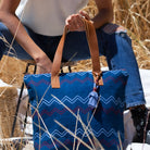 A model sits down in a wheat field and holds the Angela Tote in Lake Ripple pattern. It has horizontal zigzag stripes in red, light blue, and blue over a blue background. It has a blue tassel and leather handles.