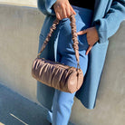 A model holding a medium cylindrical-shaped shoulder bag with a ruched body and strap against a concrete wall. 