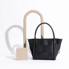 A still image of a small wide strap woven tote bag against a white wall with wood props.