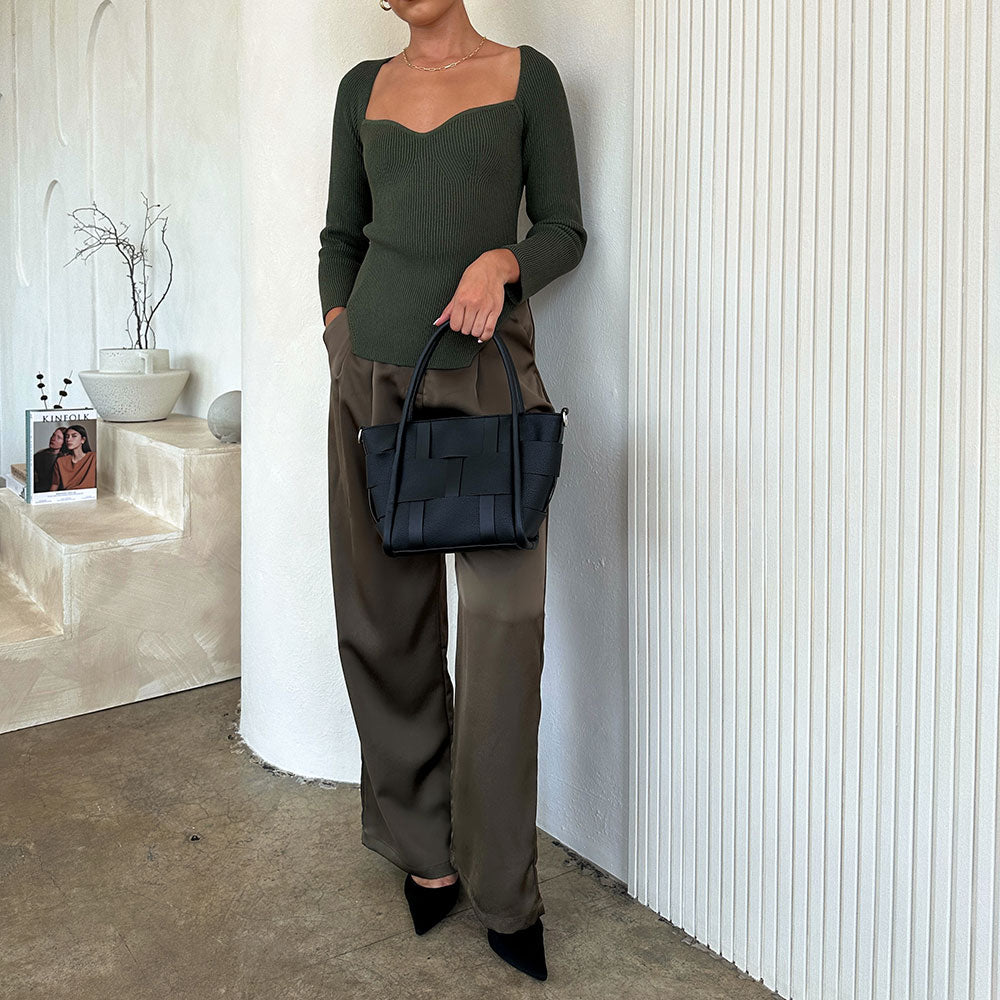 A model holding a black woven wide strap vegan leather tote bag against a white wall.