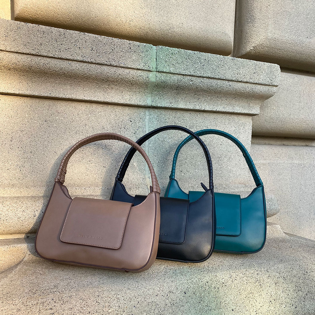 A still image of three small vegan leather shoulder bags with wrapped handle against a concrete wall. 