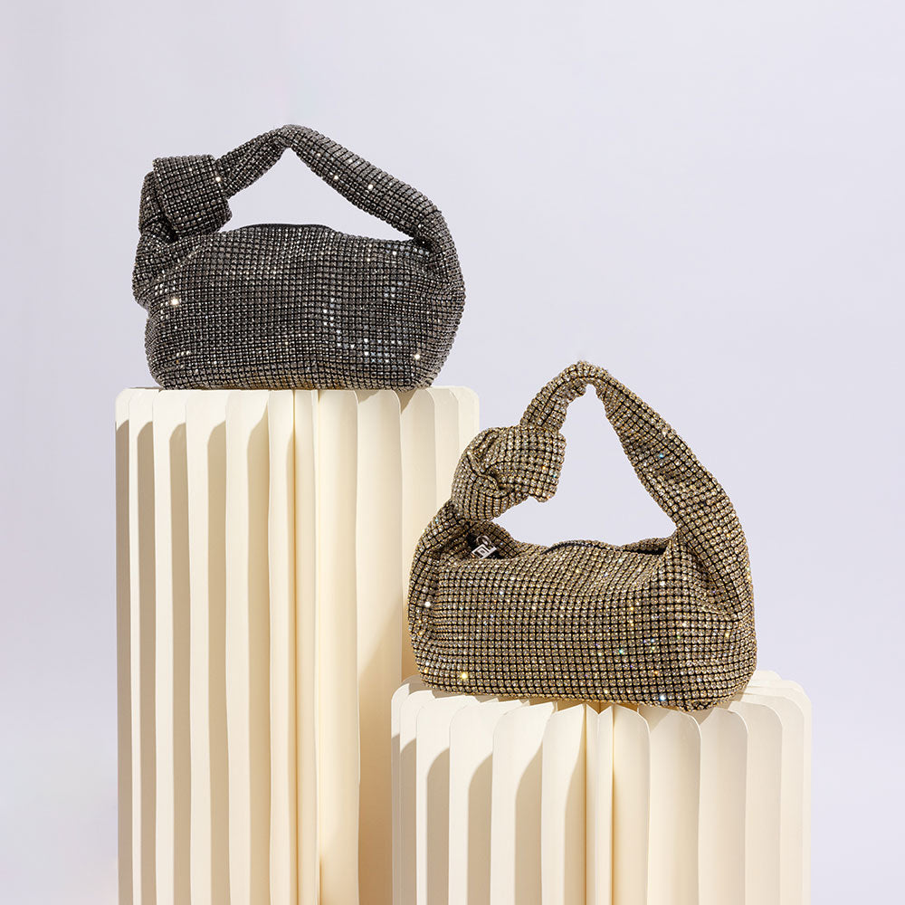 A stil image of two small crystal encrusted top handle bags with knots against a white wall. 