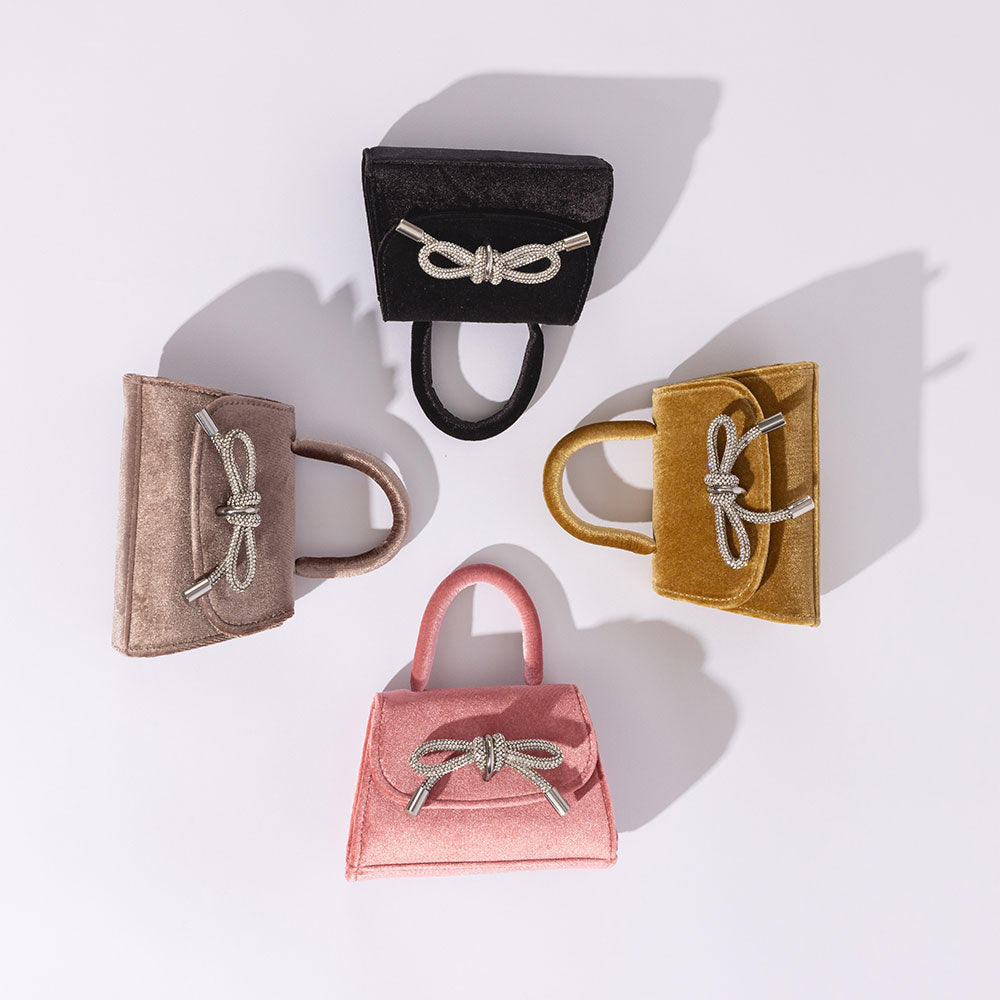 A still image of four mini velvet top handle bags with silver encrusted bow against a white wall. 