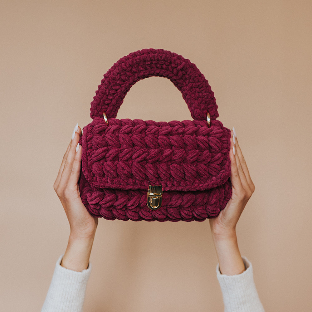 A model holding up a plum knitted crossbody handbag with gold clasps. 