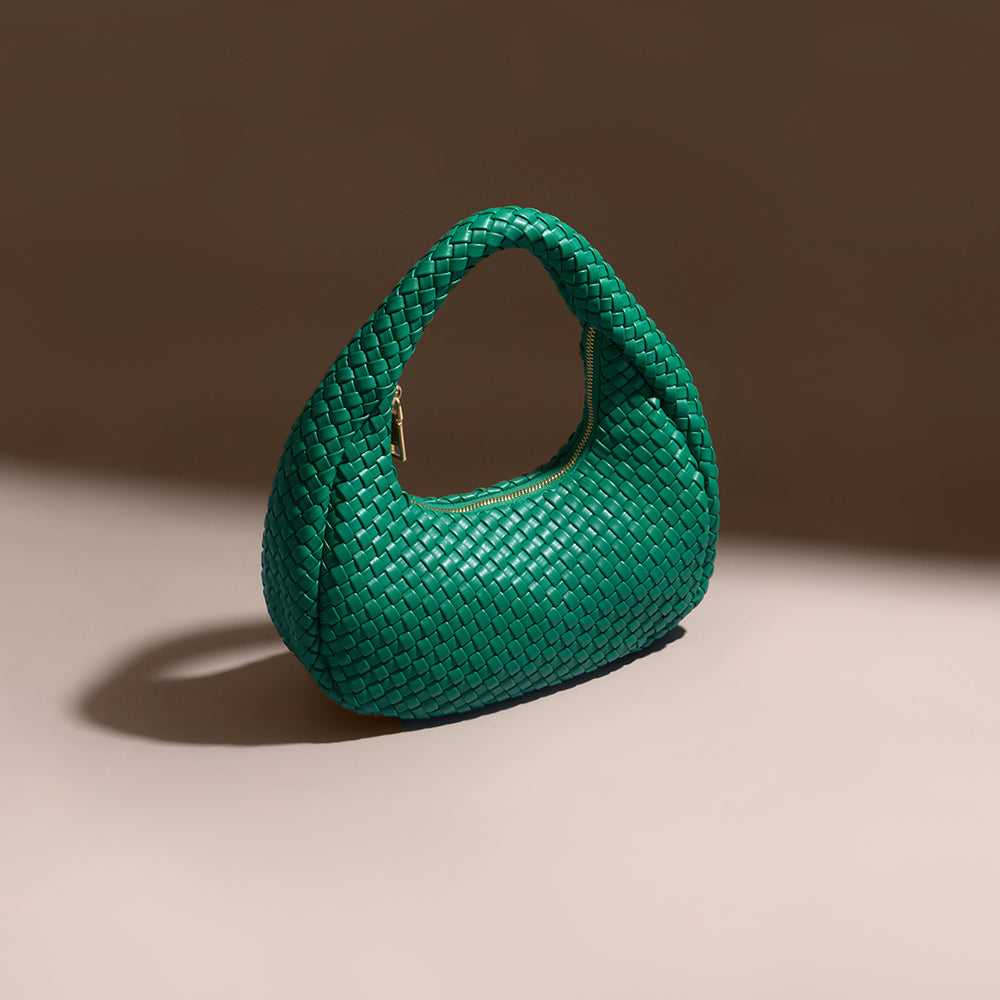 A still image of a green curved woven vegan leather shoulder bag against a brown wall. 