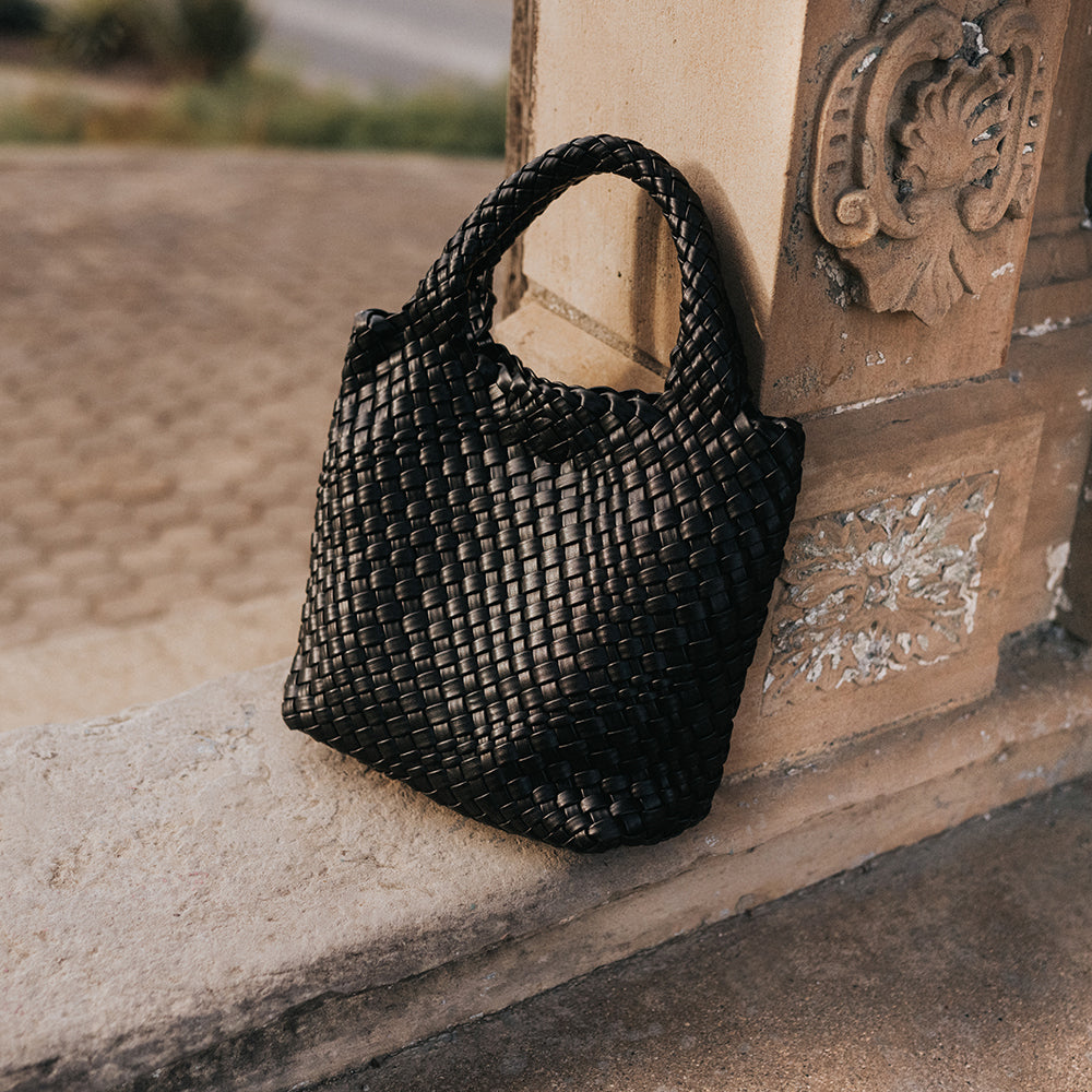 A still image of a black woven vegan leather tote bag with double handles outside against a wall. 