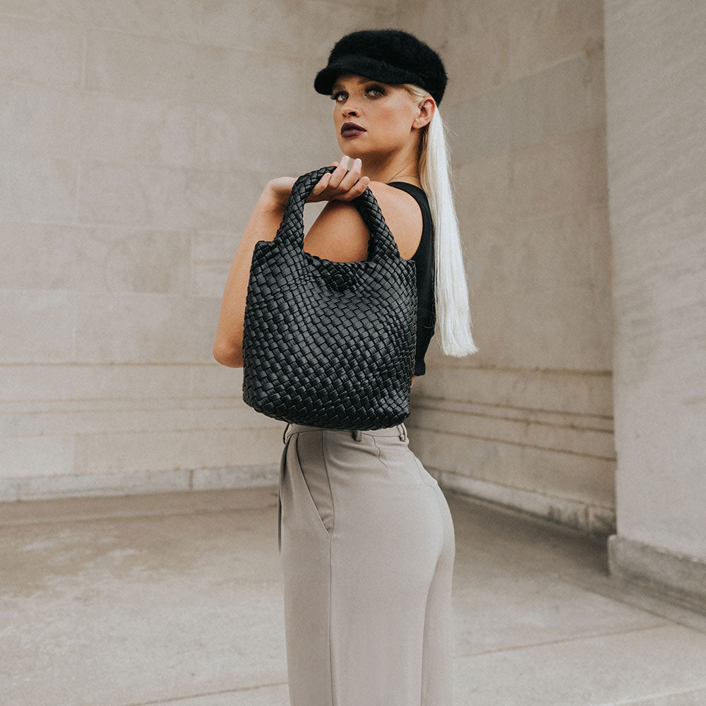 A model holding a small black woven vegan leather tote bag against a concrete wall. 