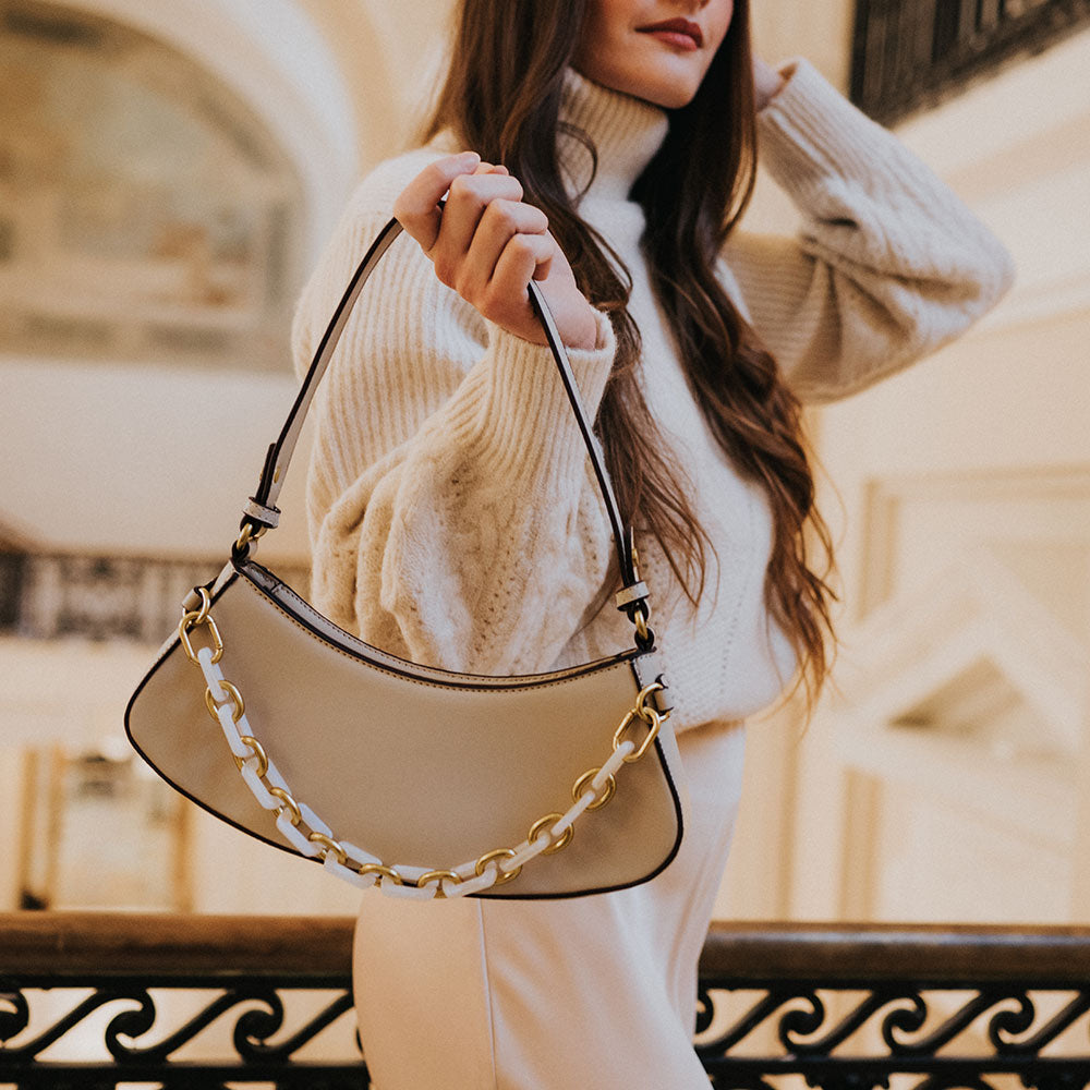 A model holding a cream recycled vegan leather shoulder bag with a chain on the front against a railing. 