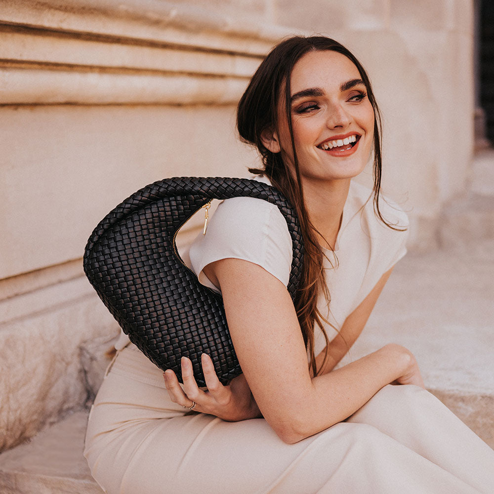 A model wearing a black curved woven vegan leather shoulder bag sitting against a wall.